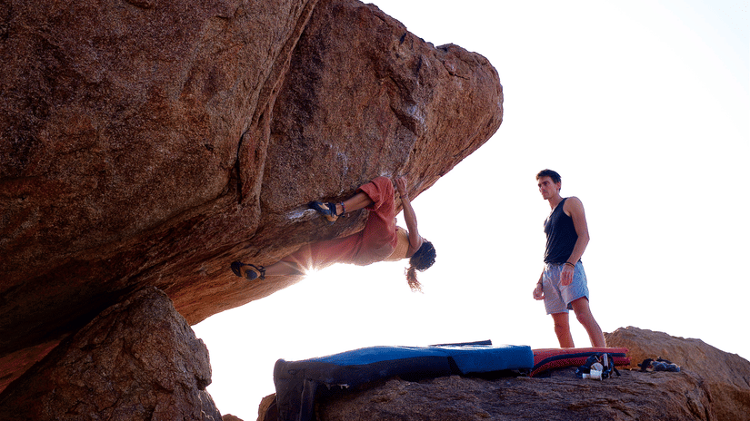 A person rock climbing on an overhang with another person standing beside them, against a bright sky