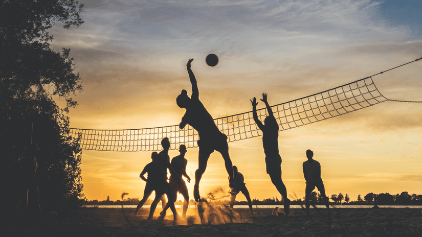 Silhouettes of people playing volleyball on a beach at sunset, with a net dividing the teams against a vibrant orange and yellow sky