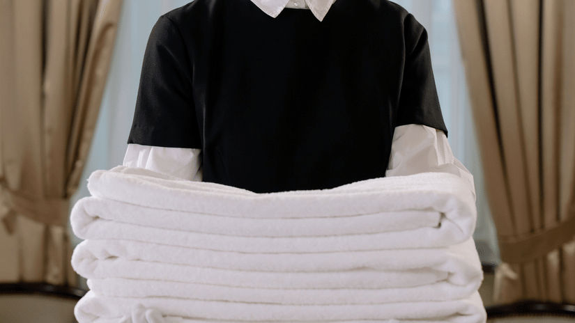 A person wearing a short-sleeved shirt and apron carefully holds a neatly folded stack of crisp white towels, possibly in a hotel or spa setting