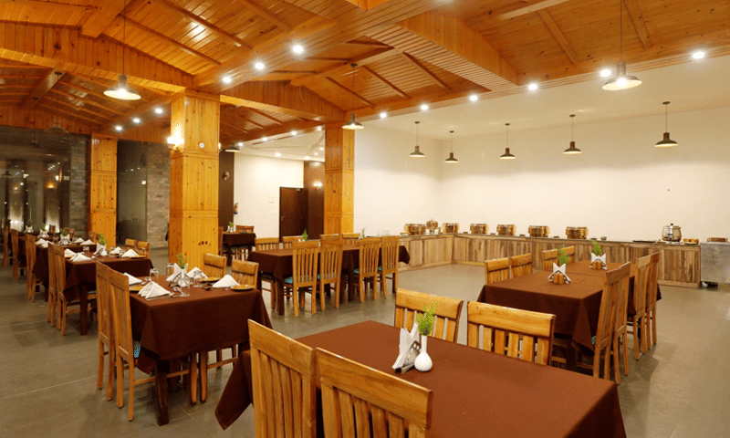A rustic, spacious dining area with wooden tables and chairs, illuminated by overhead lighting, suggesting a hotel restaurant